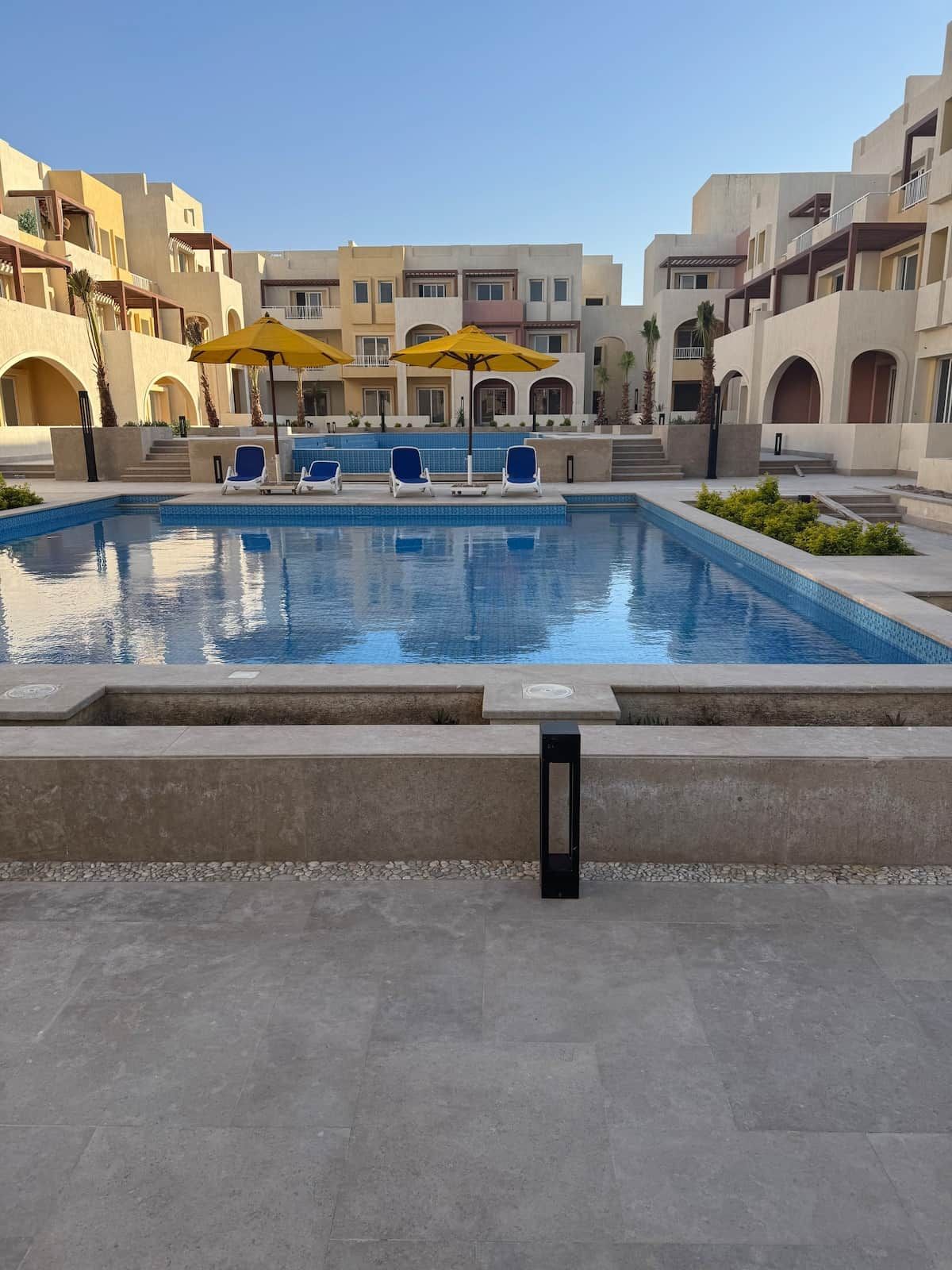 A swimming pool with lounge chairs and umbrellas, surrounded by buildings with balconies and arched doorways.