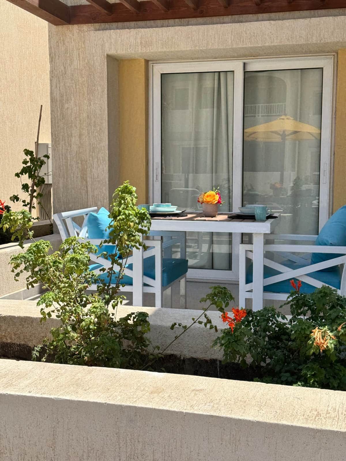 Een terras met een tafel en stoelen, een bloempot en uitzicht door een raam.