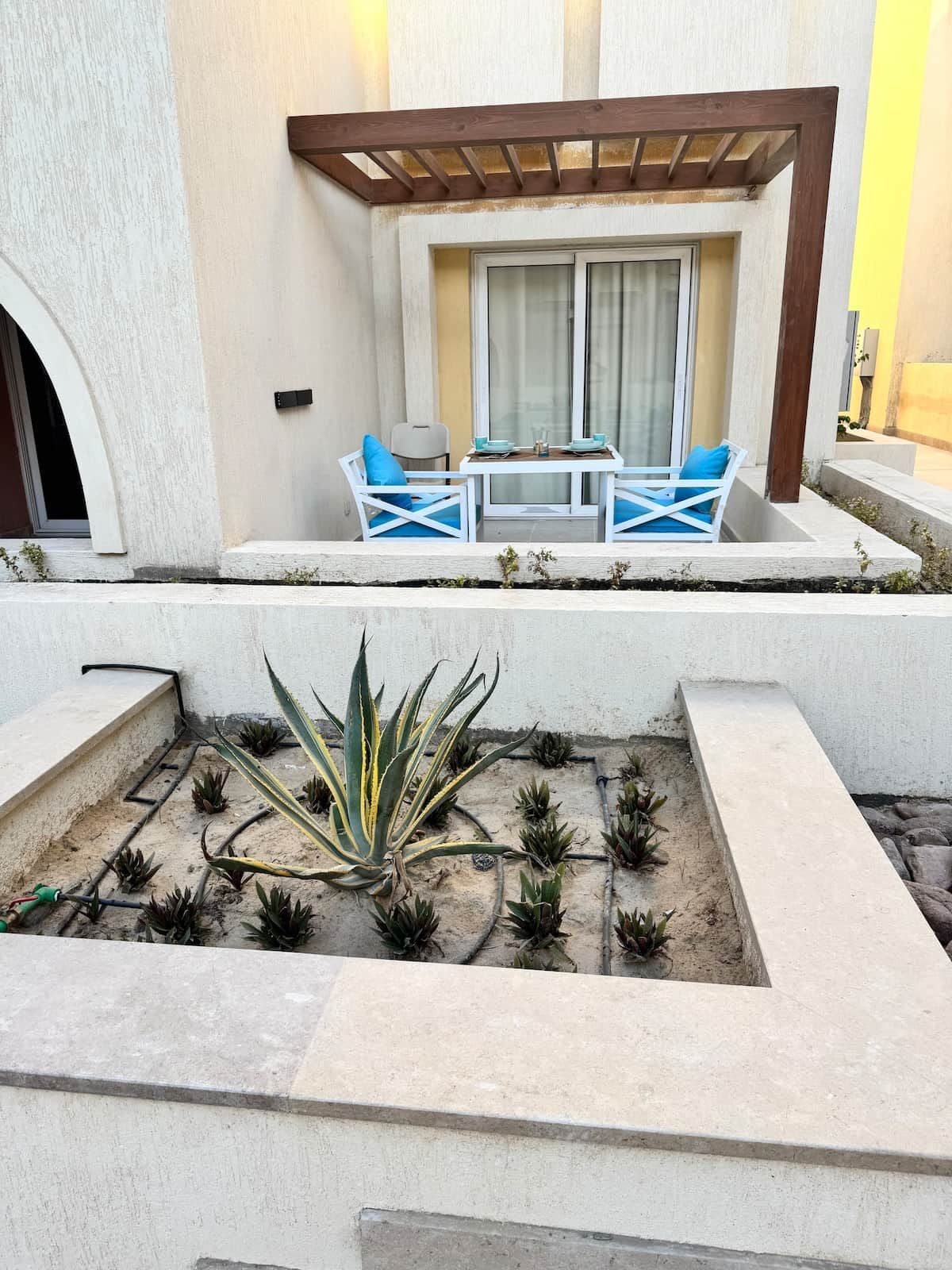 Un espacio al aire libre con mesa y sillas, una pérgola y un jardín con plantas.