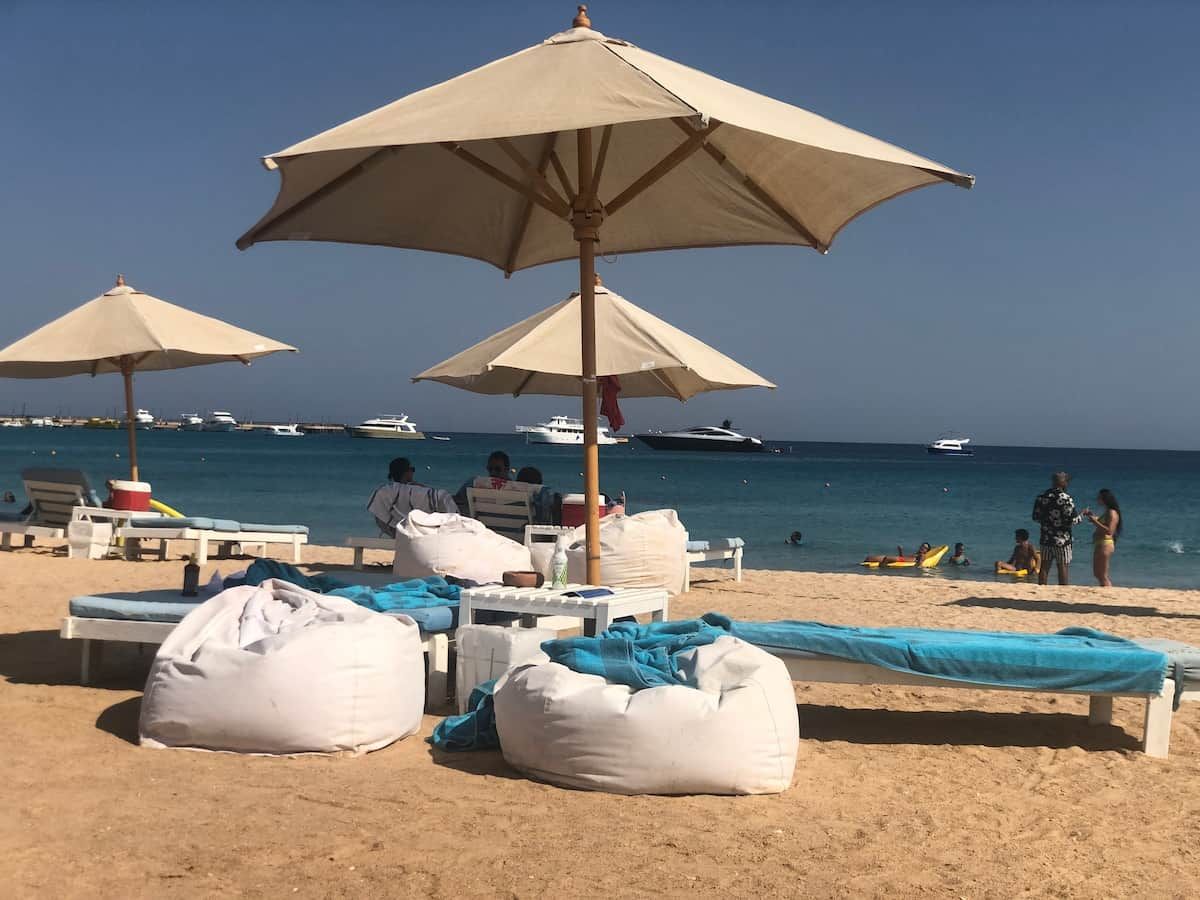 Ein Strand mit Liegestühlen, Sonnenschirmen und einem Sandbereich mit Blick auf das Meer und Boote.