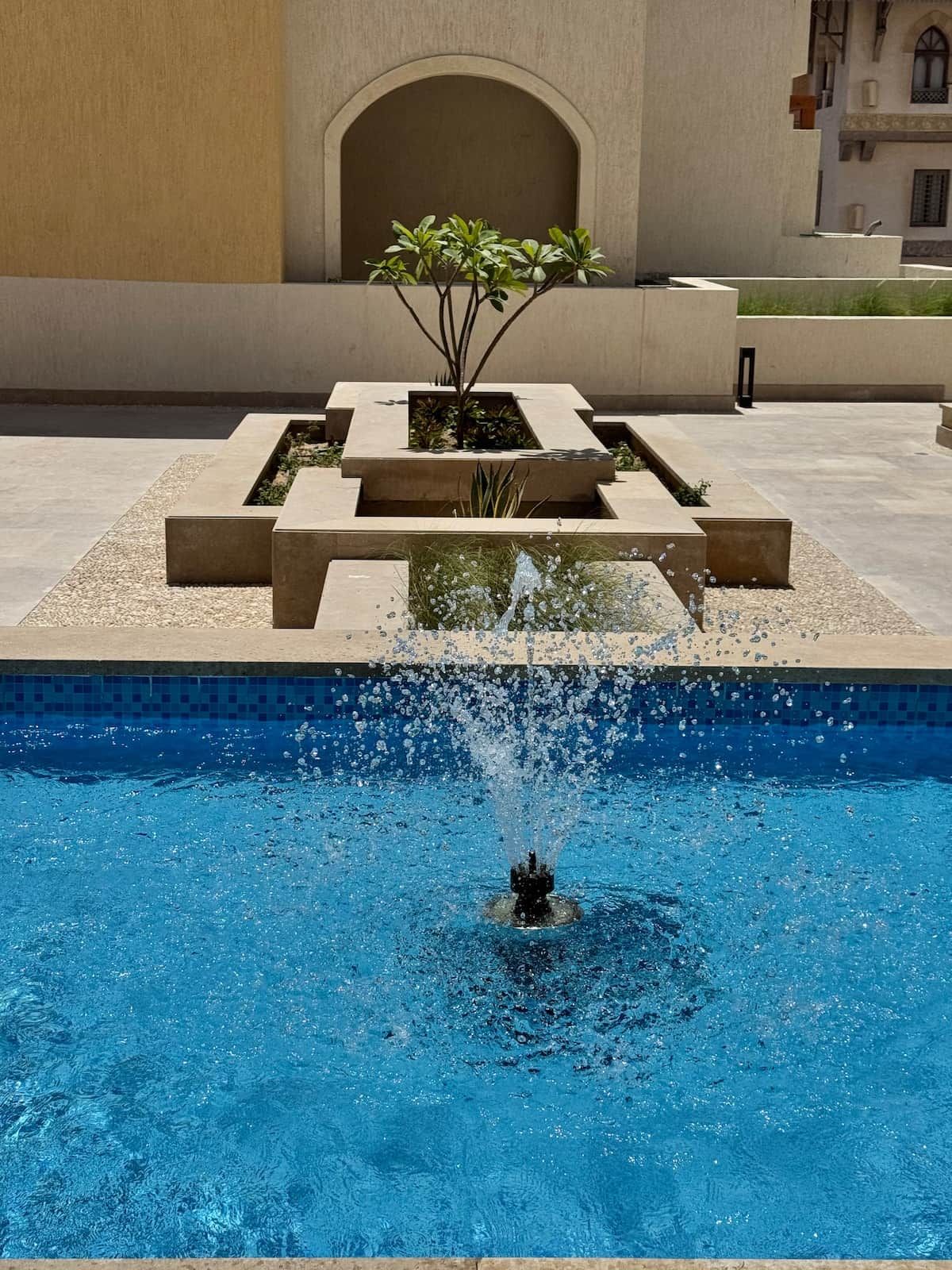 Ein Schwimmbecken mit Springbrunnen, umgeben von einem geometrisch gestalteten Pflanzgefäß mit Pflanzen.
