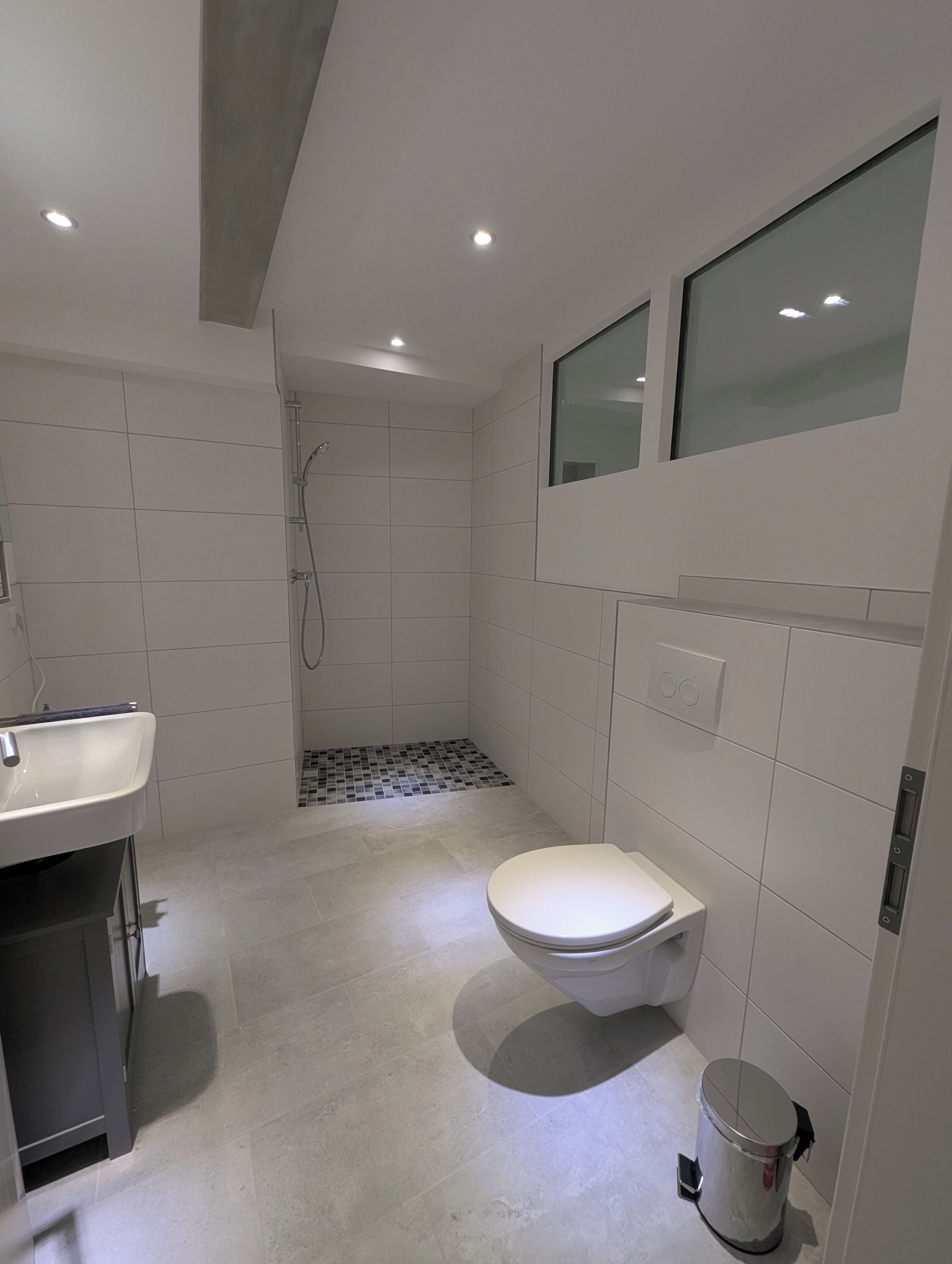 Een moderne badkamer met toilet, wastafel, douche en een klein prullenbakje.
