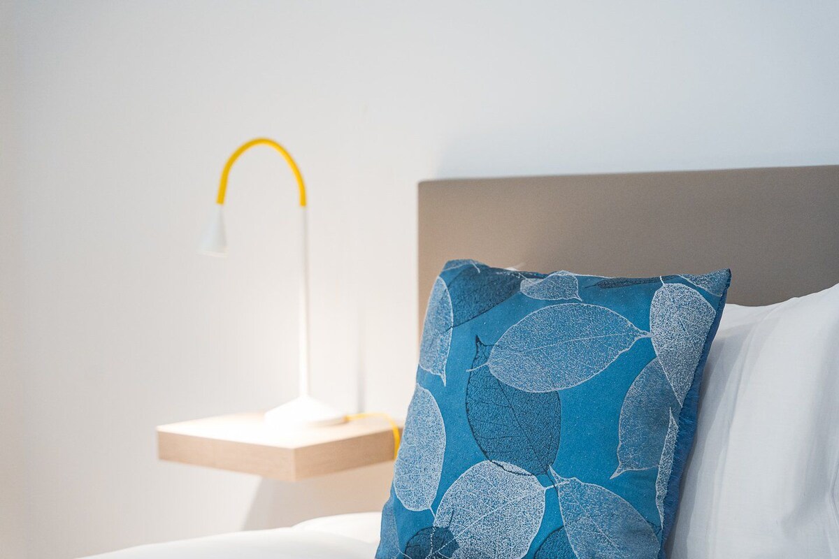 Cama bem arrumada com travesseiro branco e travesseiro azul com desenhos de folhas, mesa de cabeceira com abajur branco e parede lisa.