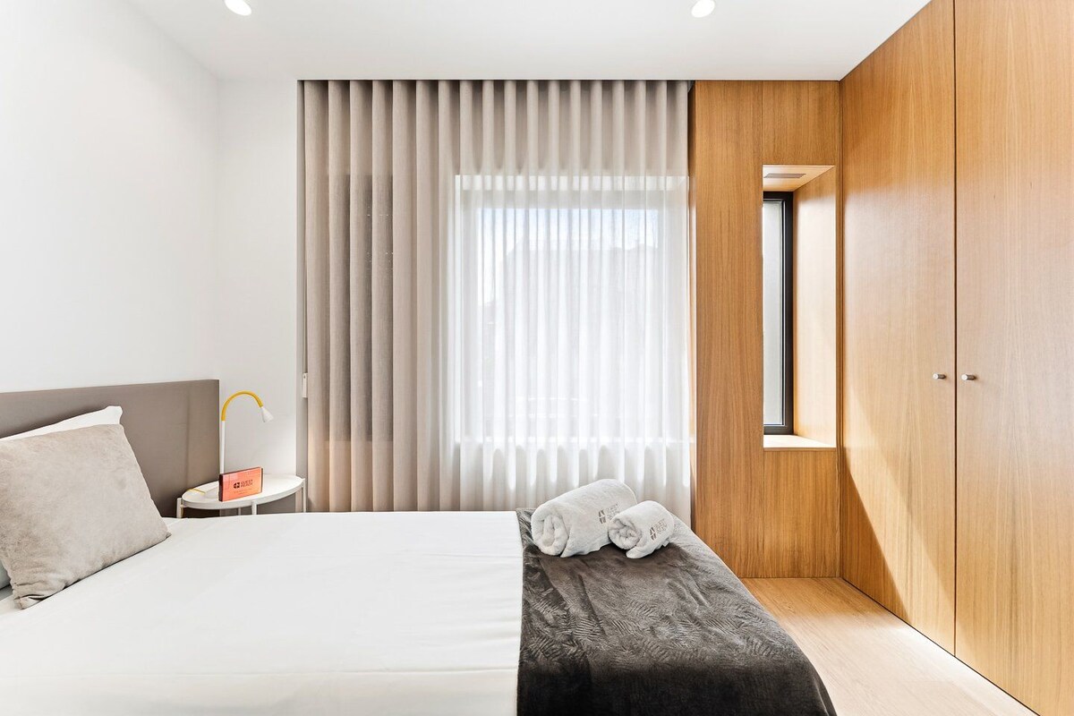 A modern hotel room with a bed, a bedside table, a nightstand, a window with curtains, and a wardrobe.