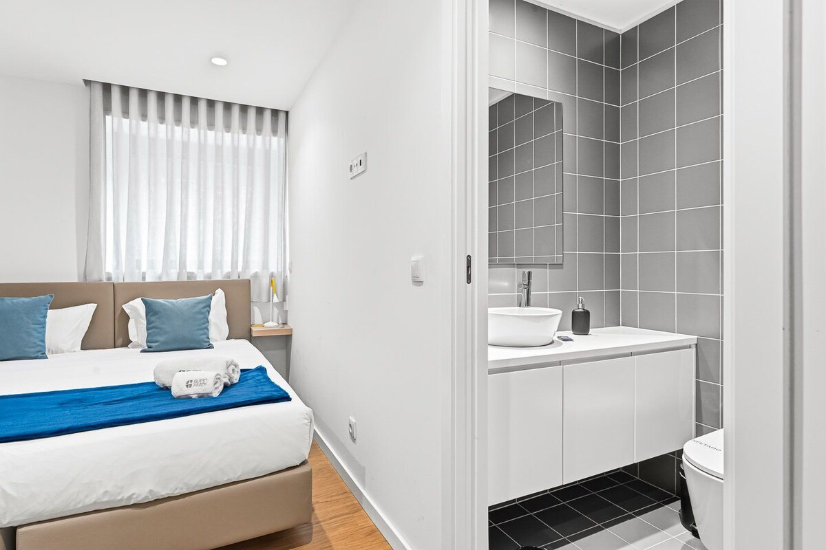 A modern hotel room with a bed, a bedside table, a mirror, and a bathroom with a sink and toilet.