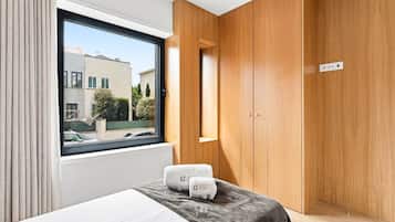 Un dormitorio moderno con un ventanal grande, un armario de madera y una cama con una manta gris y dos toallas.
