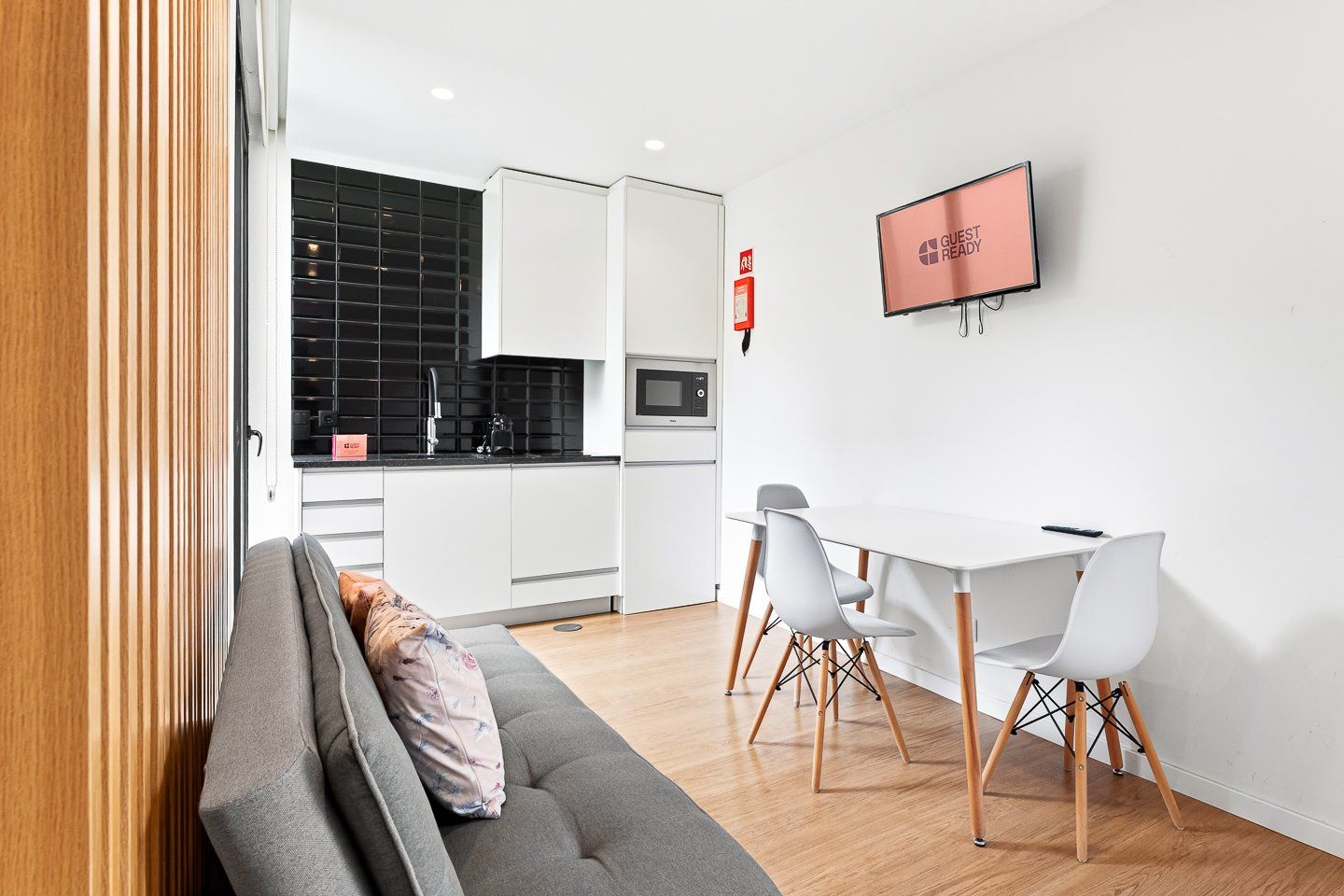 A modern kitchen with white cabinets, a black backsplash, and a built-in oven. There is a dining area with a white table and chairs, and a TV mounted on the wall.