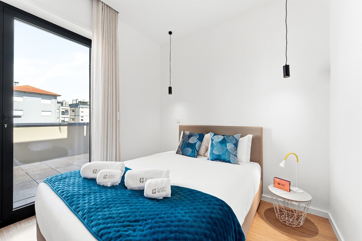 A hotel room with a bed, a bedside table with a lamp, a view of the city, and a balcony.