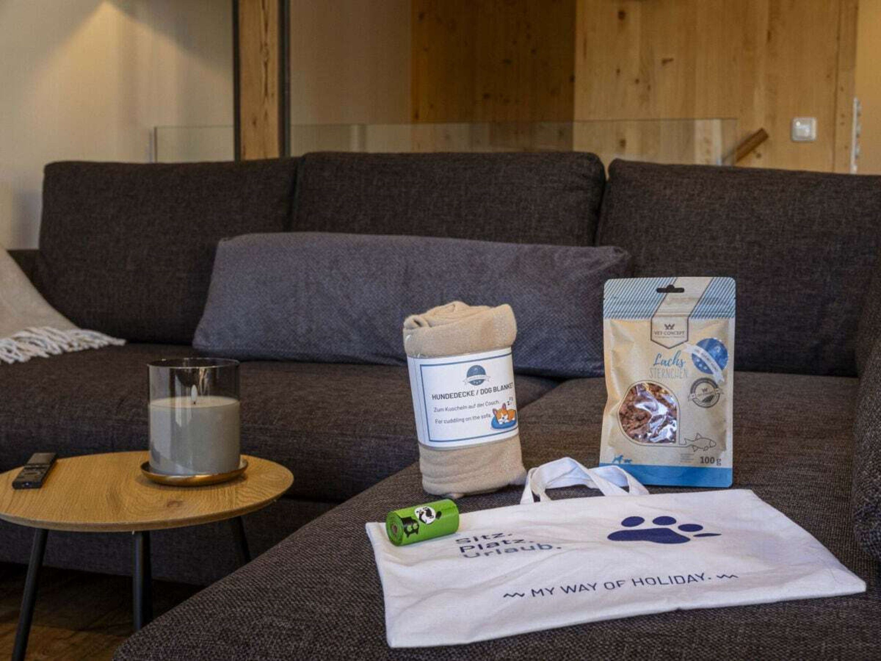 Ein Wohnzimmer mit einem grauen Sofa, einem Glaswindlicht, einer aufgerollten Decke, einem Futtersack für Haustiere und einer Stofftasche mit Pfotenabdruck.