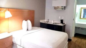 A hotel room with a bed, bedside table, lamp, and a kitchenette area with a sink and countertop. - 1 King Bed | Mob Acc RI Shwr Nsmk Micfridg (Savannah GA)