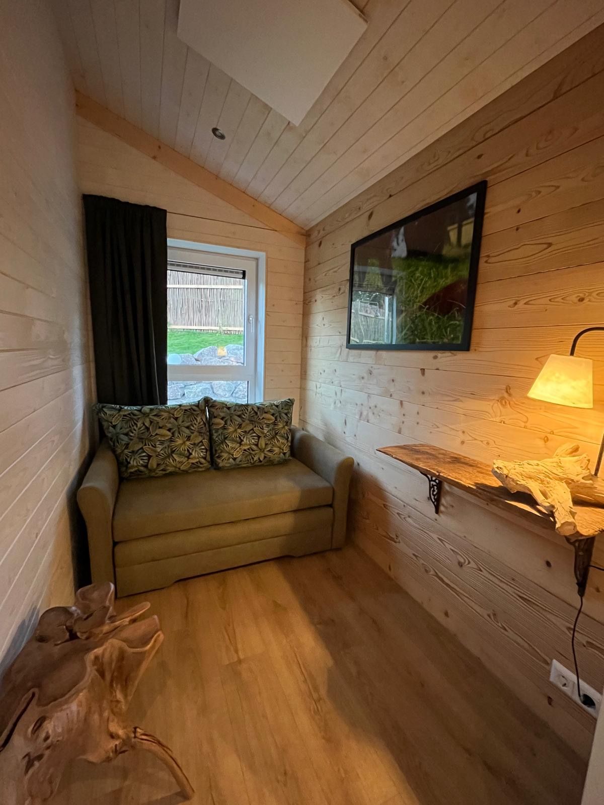 A cozy room with a wooden ceiling, a window with a view of a grassy area, a sofa with patterned cushions, and a wall-mounted lamp.