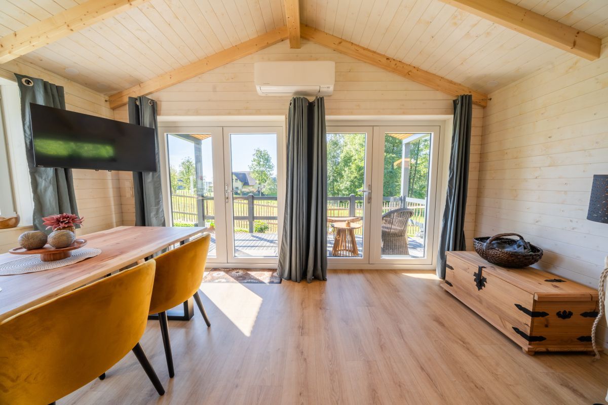 A wooden interior with a dining table, chairs, a TV, and a view through large windows.
