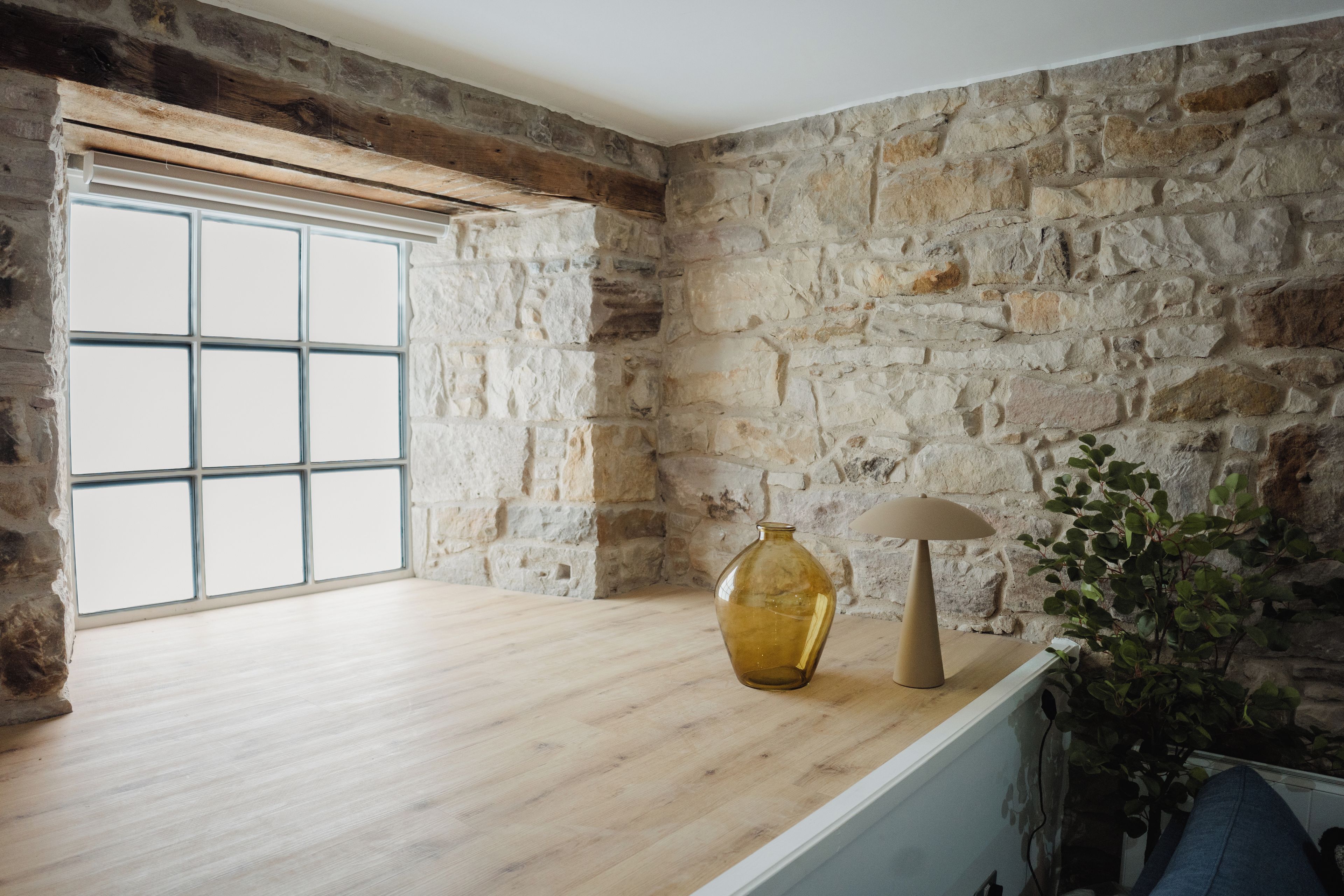 Una habitación con pared de piedra, un ventanal grande, suelo de madera, un jarrón, una lámpara y una planta.