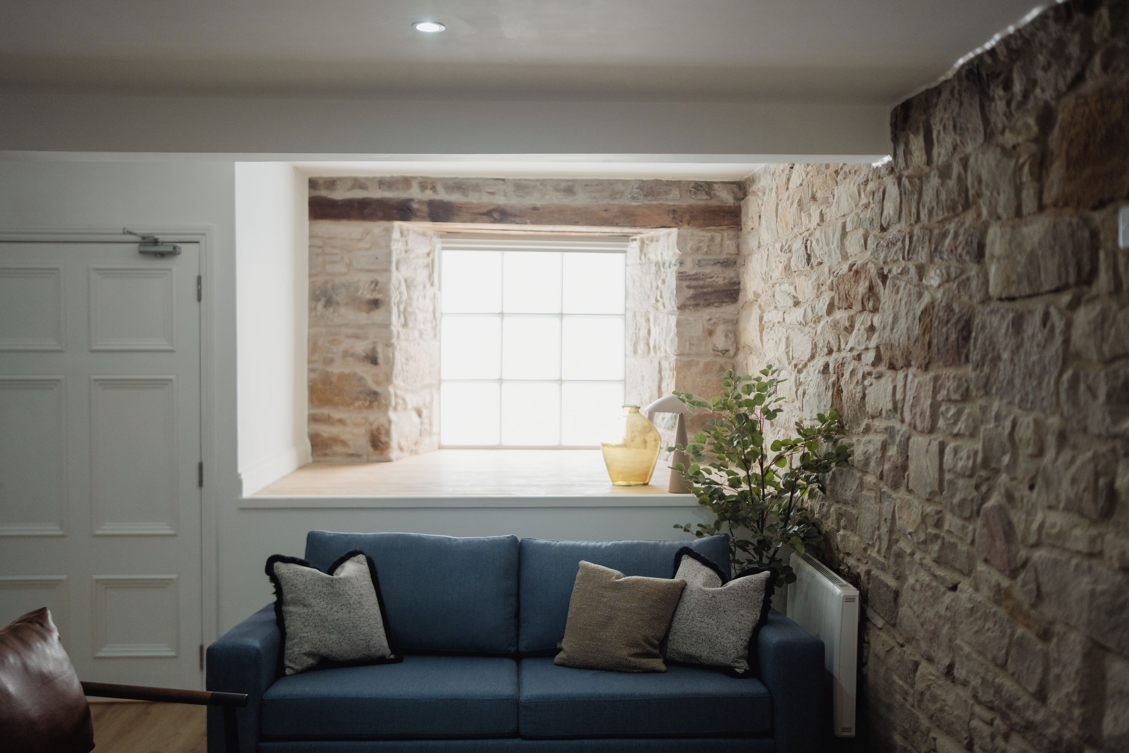 Una habitación con pared de piedra, ventana con vistas, sofá azul con cojines y una planta.