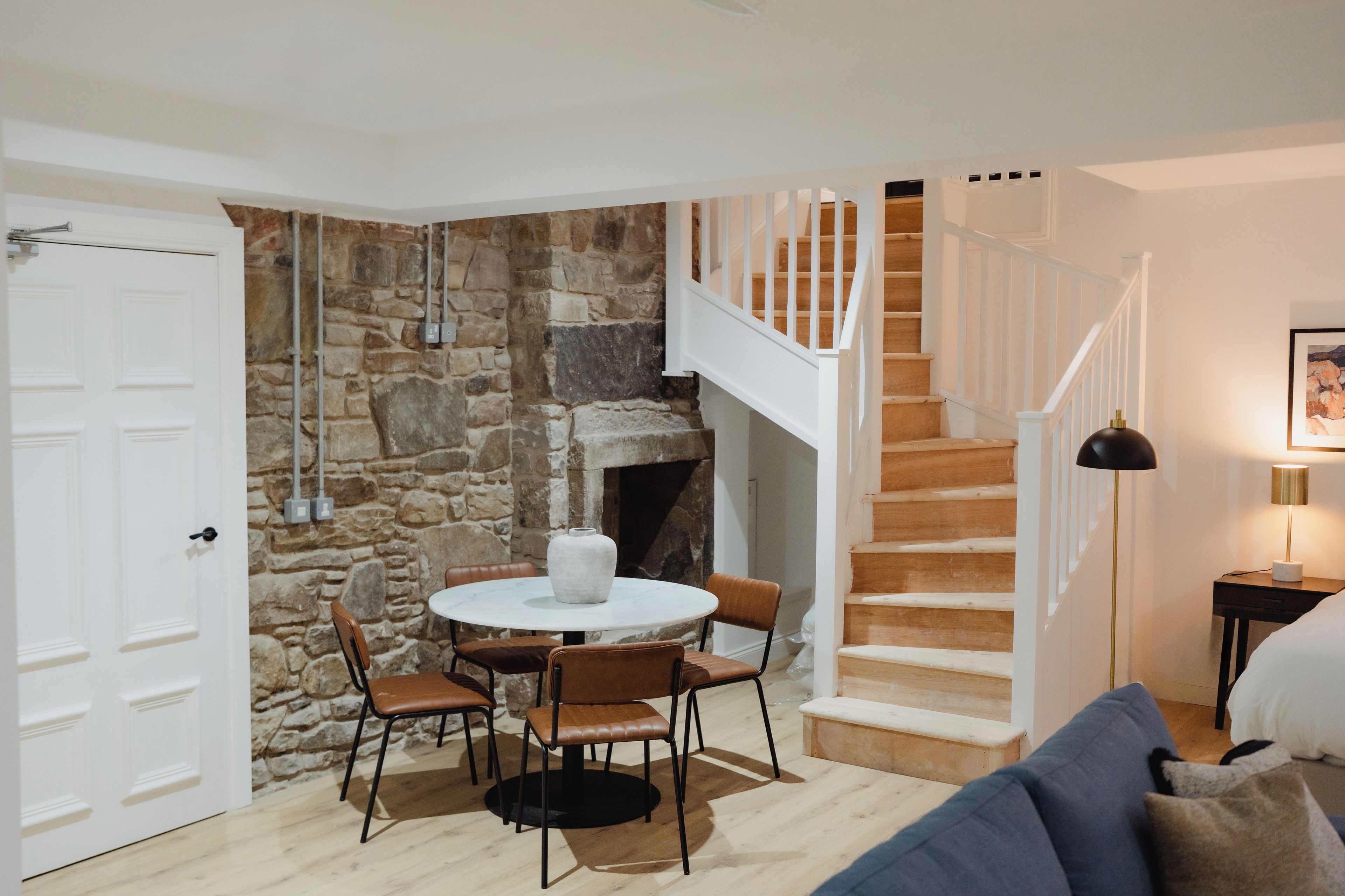Una habitación acogedora con pared de piedra, chimenea, una mesa redonda con sillas y una escalera con barandillas blancas.