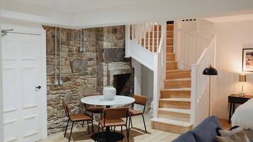 Una habitación acogedora con pared de piedra, chimenea, una mesa redonda con sillas y una escalera con barandillas blancas.