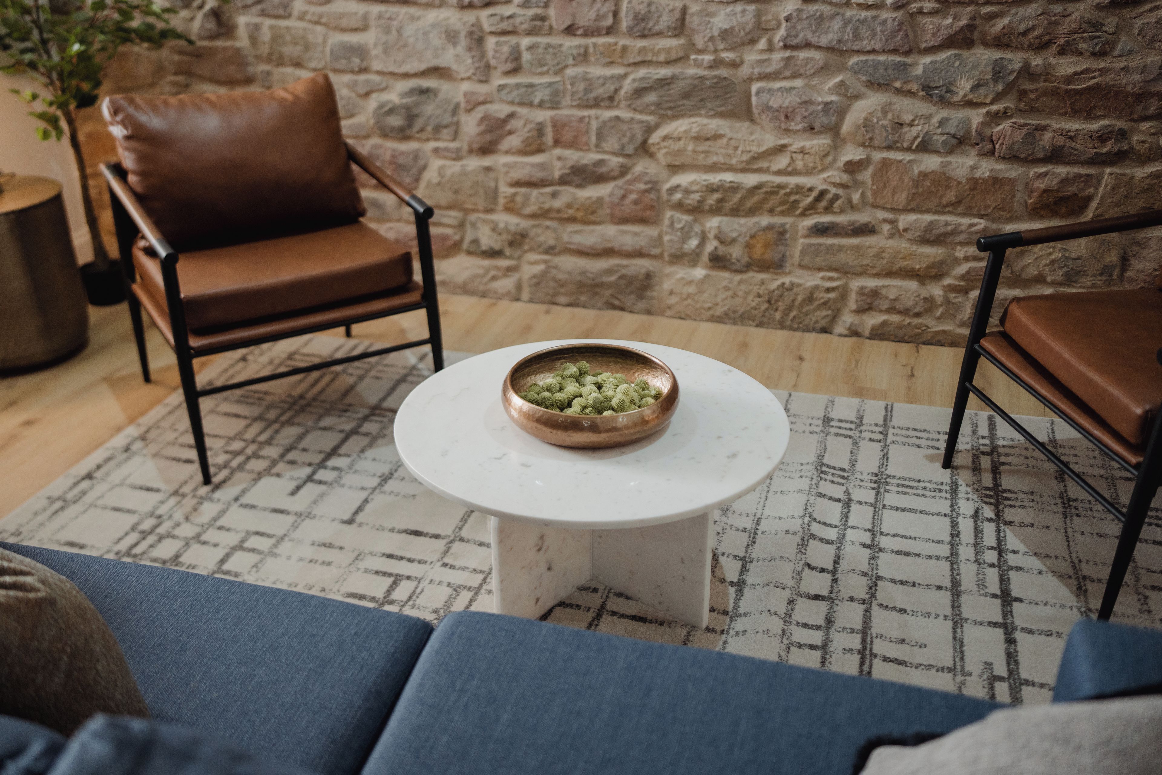 A room with a stone wall, a brown leather chair, a white coffee table with a copper bowl, and a blue sofa.