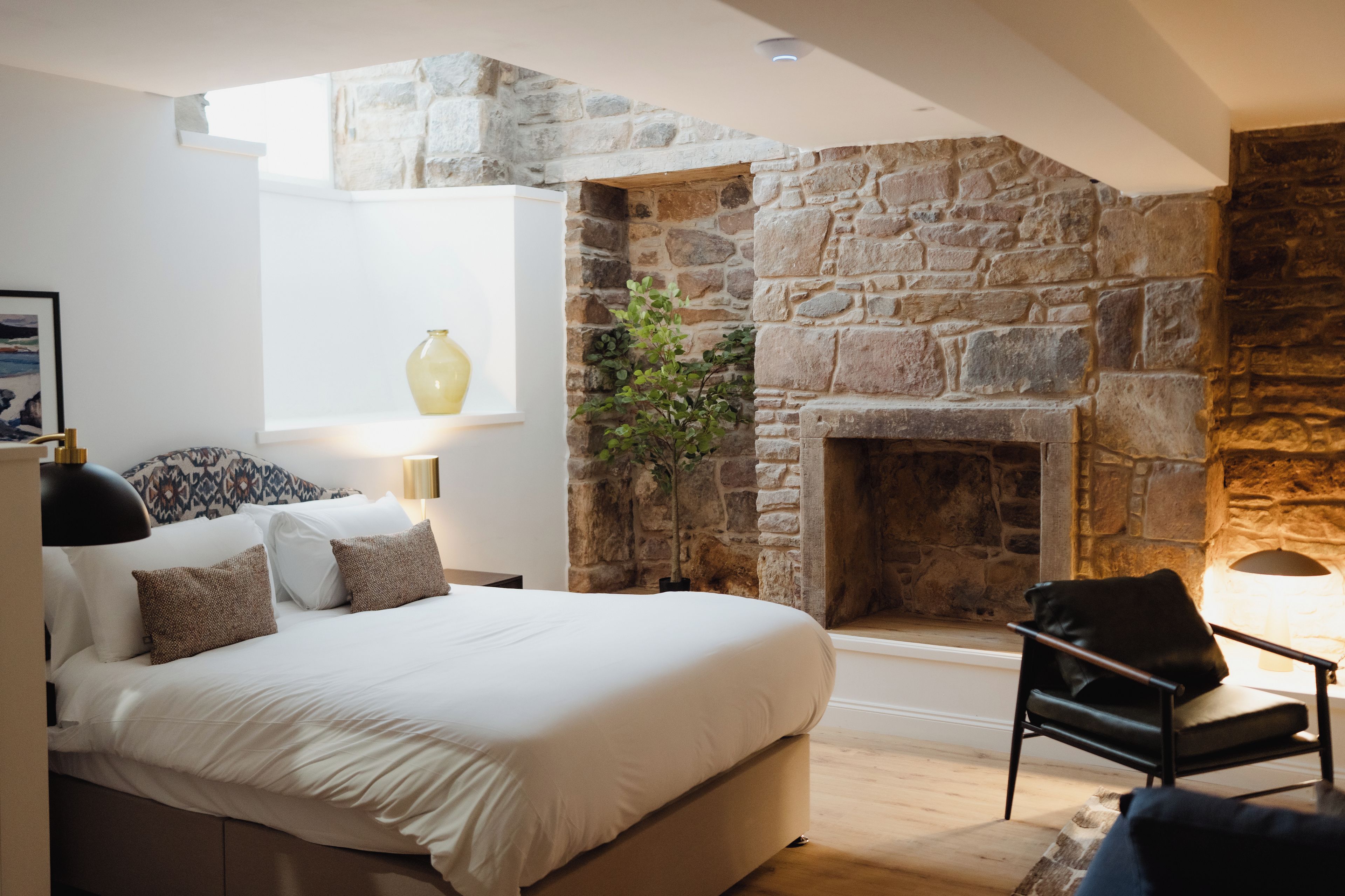 A bedroom with a stone fireplace, a bed with white bedding, a side table with a lamp, and a window with a vase.