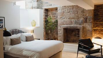 A bedroom with a stone fireplace, a bed with white bedding, a side table with a lamp, and a window with a vase.
