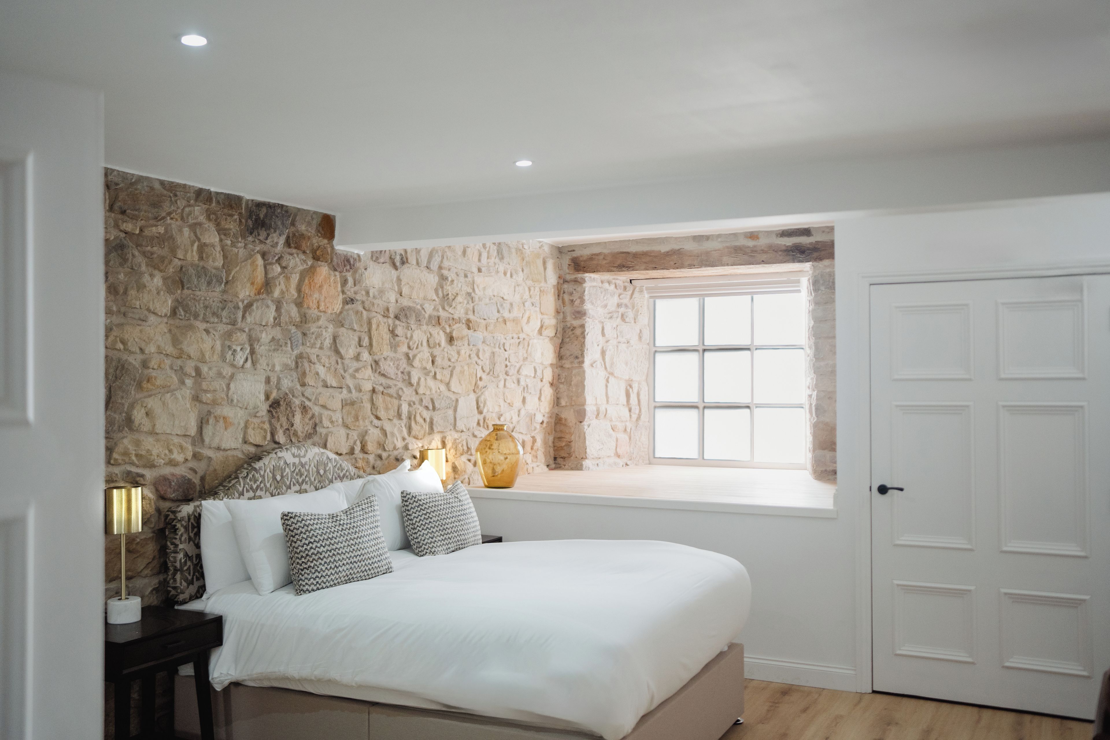 A bedroom with a stone wall, a bed with white bedding, a window, and a door.
