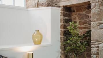 A bedroom with a stone wall, a bed, a nightstand, a lamp, a vase, and a plant.