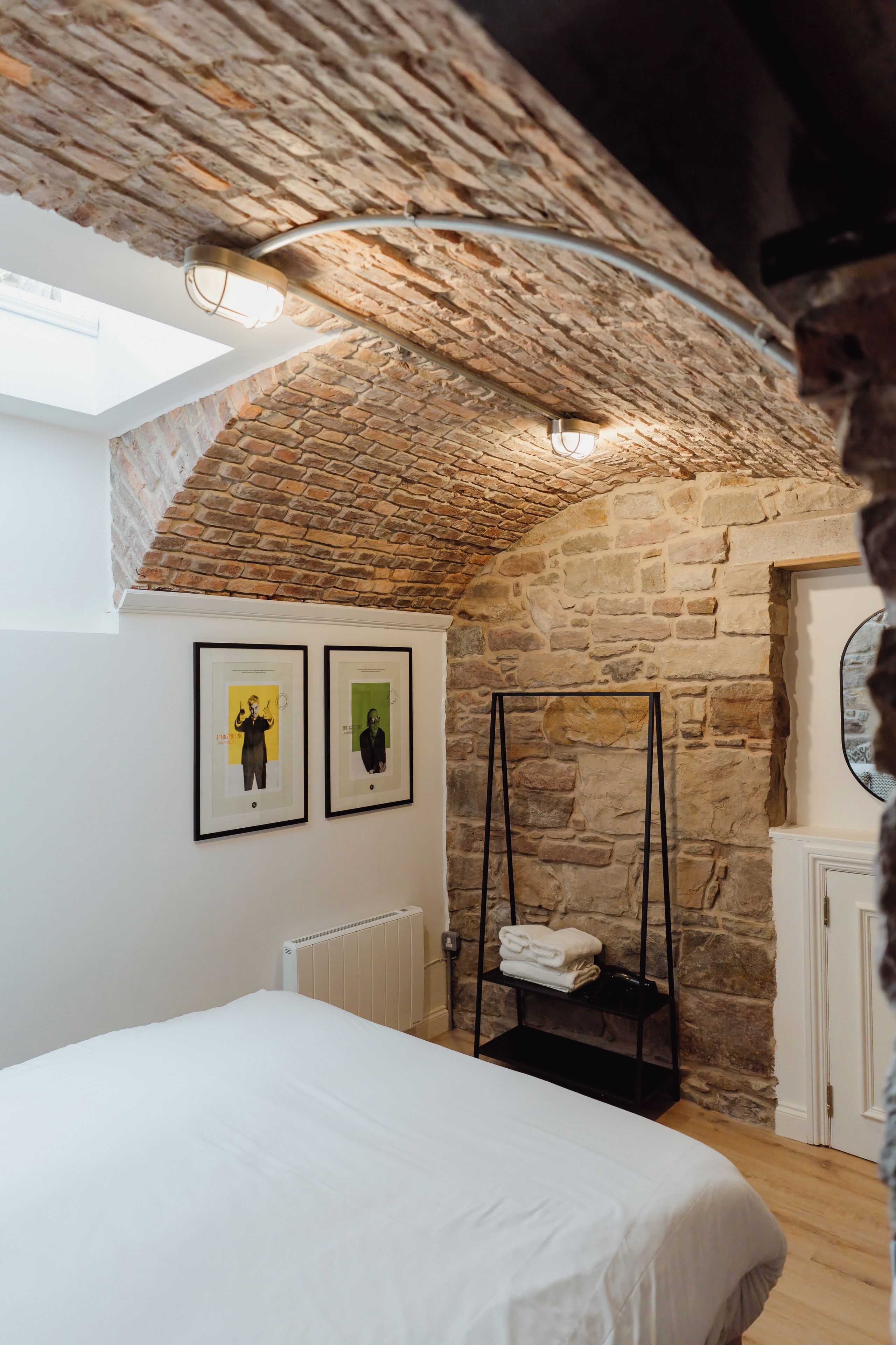 Una camera da letto con soffitto spiovente, pareti in mattoni a vista, un letto, un termosifone e due quadri appesi al muro.