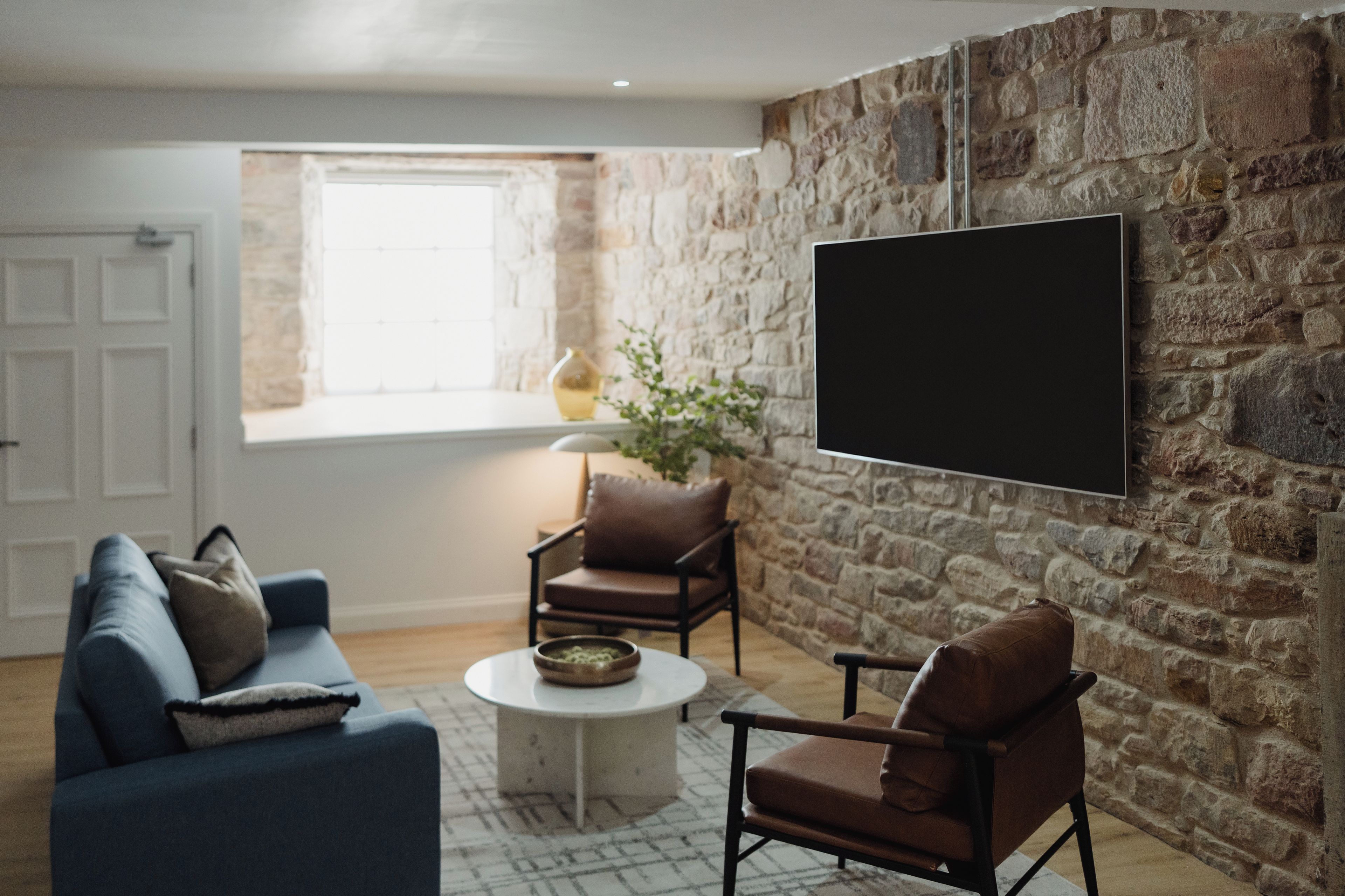 Ein Wohnzimmer mit Steinwand, einem Wand-Fernseher, einem blauen Sofa, zwei Sesseln, einem Couchtisch und einem Fenster mit Aussicht.