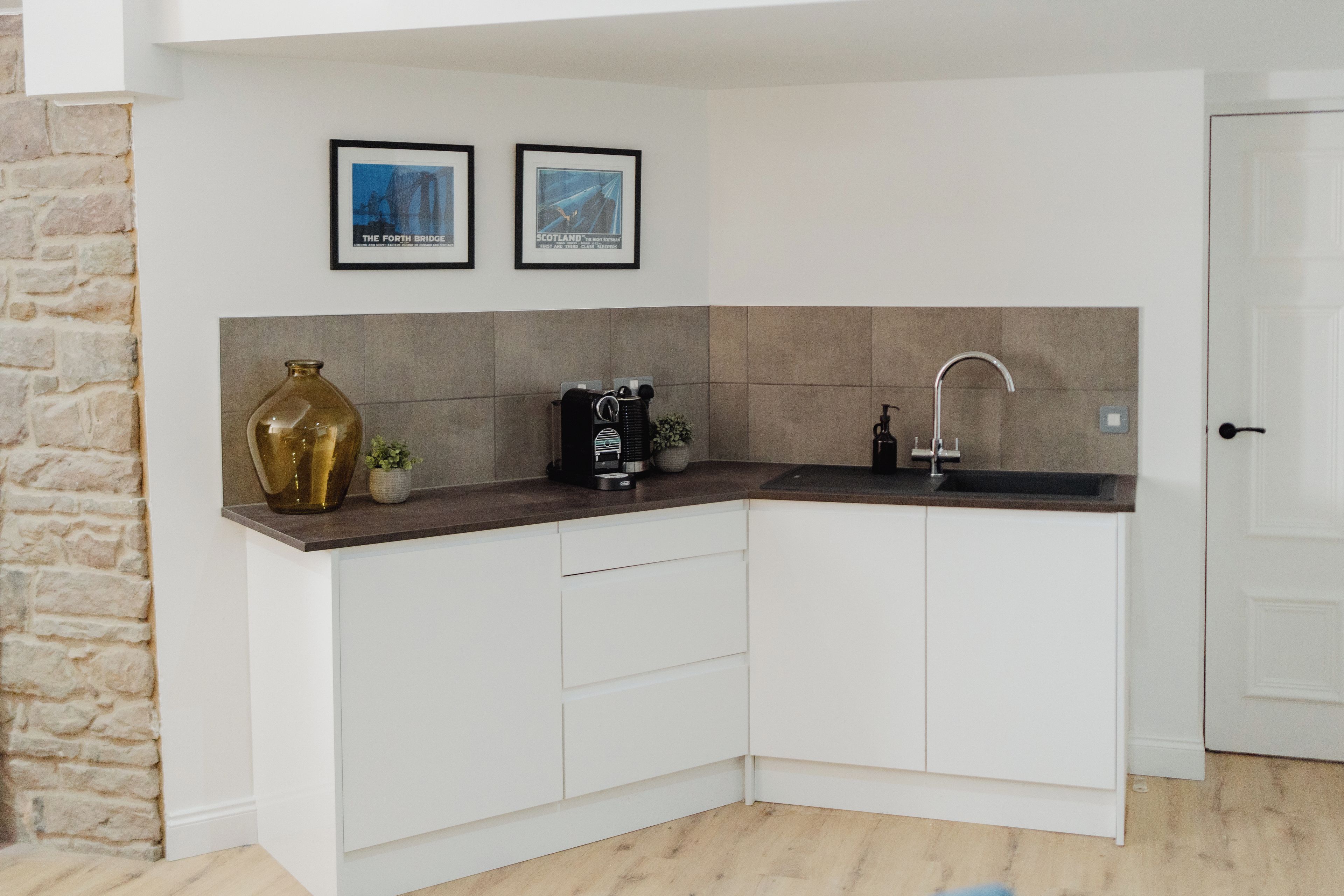 A corner kitchen with white cabinets, a coffee machine, a vase, and two framed pictures on the wall.