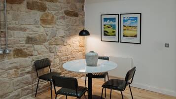 Een kamer met een stenen muur, een ronde tafel met stoelen, een vaas, en twee ingelijste foto's aan de muur.