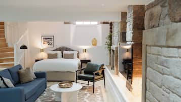 A bedroom with a bed, a blue sofa, a coffee table, and a chair. There is a staircase on the left and a stone wall on the right.