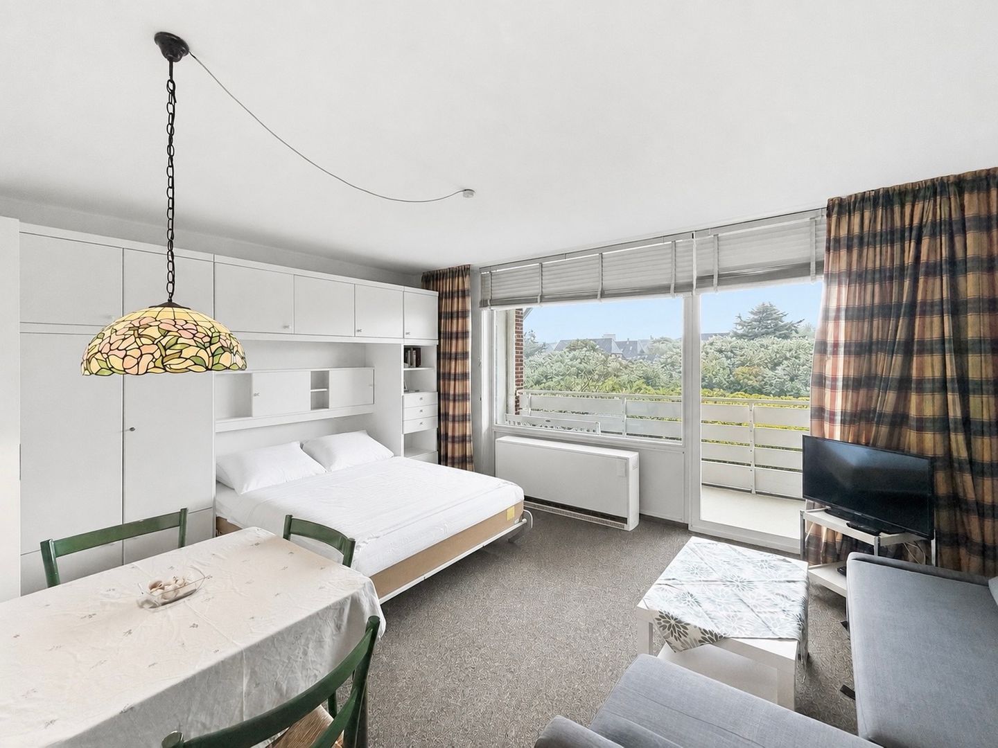 Ein modernes Hotelzimmer mit einem Bett, einem Esstisch, einem Fernseher und einem großen Fenster mit Vorhängen.