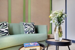 A modern living room with a green sofa, patterned pillows, a small round table with a vase of flowers, and a stack of books. - One-Bedroom Superior Apartment (Paris)
