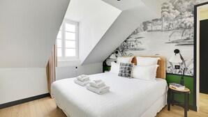 A bedroom with a sloped ceiling, a bed with white linens, a mural of a savannah scene, and a small table with books and a vase. - One-Bedroom Superior Apartment (Paris)