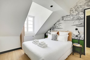 A bedroom with a sloped ceiling, a bed with white linens, a mural of a savannah scene, and a small table with books and a vase. - One-Bedroom Superior Apartment (Paris)