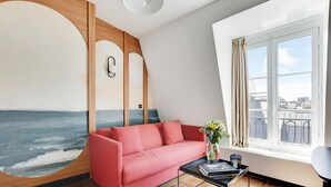 A modern living room with a red sofa, a coffee table with books, a wall mural, and a window with a city view. - One-Bedroom Superior Apartment (Paris)
