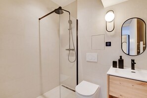 A modern bathroom with a glass-enclosed shower, a white toilet, a wooden vanity with a mirror, and a wall-mounted light fixture. - One-Bedroom Superior Apartment (Paris)