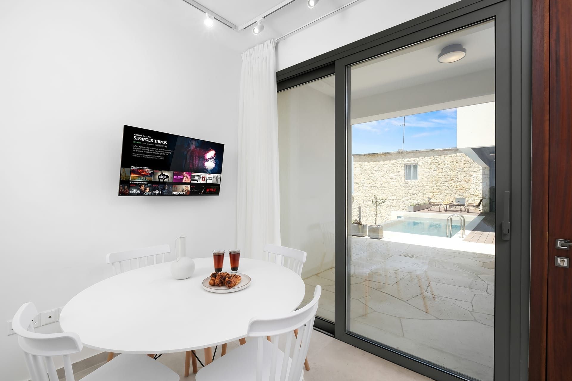 Una habitación moderna con una mesa y sillas blancas, una puerta de vidrio que da a un área exterior con piscina y pared de piedra.