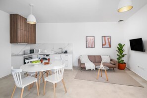 Una cocina moderna con armarios blancos, una mesa de comedor con sillas, un sofá, un televisor montado en la pared y una planta en maceta.