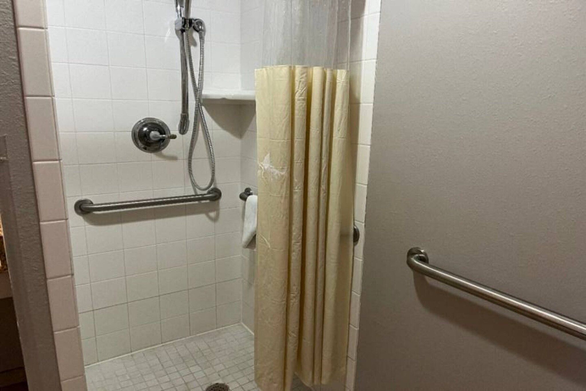 Un baño con paredes de azulejos blancos, una ducha con cabezal de ducha manual, un perchero para toallas y una puerta con picaporte de metal.