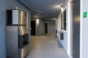 A corridor with a stainless steel refrigerator and a water dispenser on the left, a door with a green sign on the right, and a hallway leading to another room. - 2 Queen Beds | Suite, Non-Smoking, Micfridge (Ridgecrest CA)
