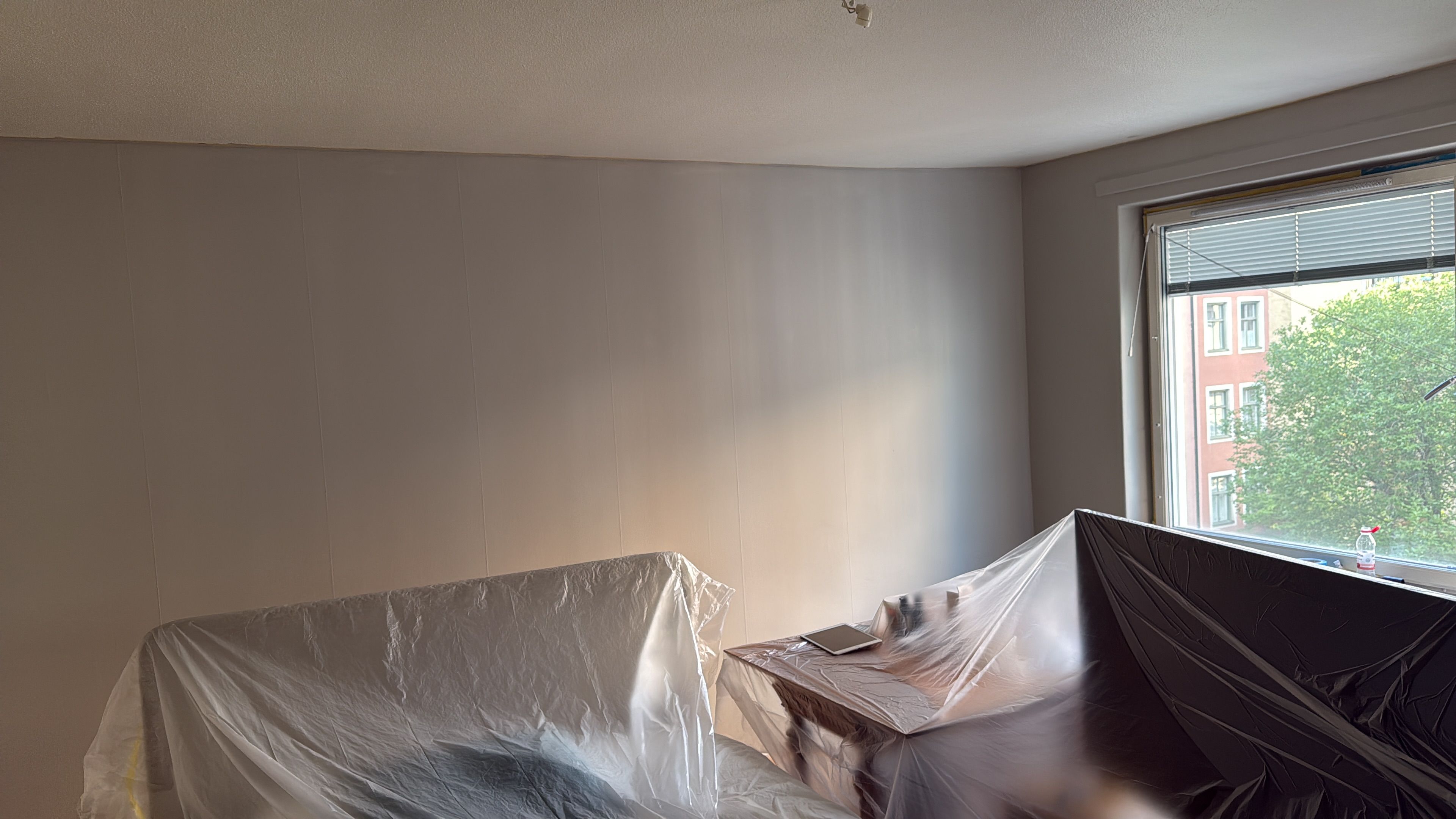 A room with a large window, a bed covered in plastic, and a view of buildings outside.