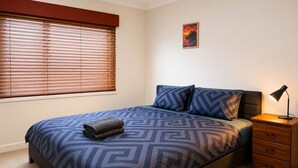Un dormitorio con cama, mesita de noche, lámpara, ventana con persianas y un cuadro colgado en la pared.