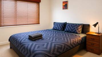 A bedroom with a bed, a nightstand, a lamp, a window with blinds, and a wall-mounted picture.
