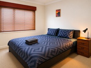 A bedroom with a bed, a nightstand, a lamp, a window with blinds, and a wall-mounted picture. - Spacious Bali Retreat | Gaming, Gym & Pool access (Tarneit)