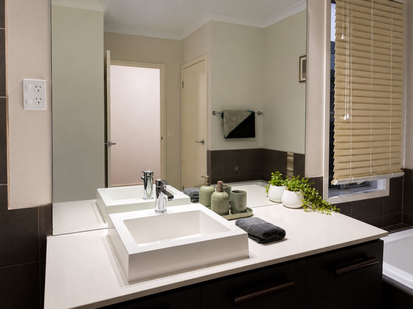 A modern bathroom with two sinks, a mirror, a towel rack, and a window with blinds.