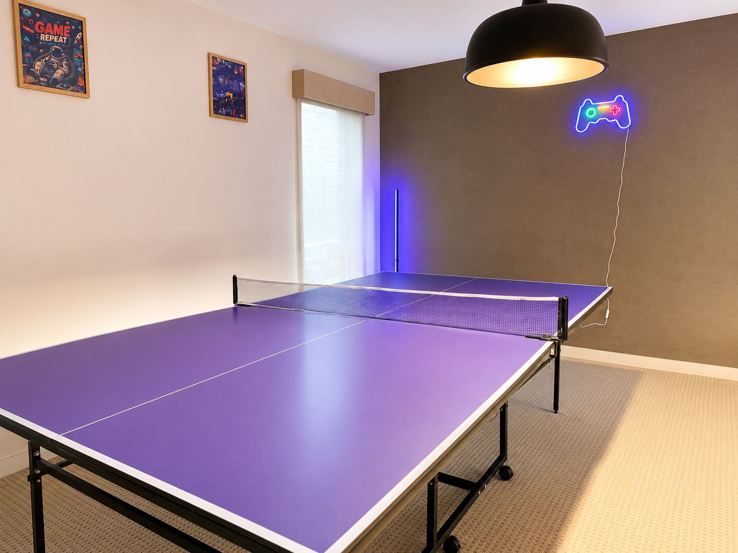 A room with a ping pong table, a neon game controller sign, and two posters on the wall.