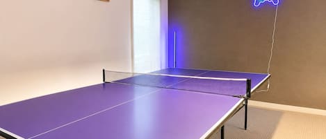 A room with a ping pong table, a neon game controller sign, and two posters on the wall.