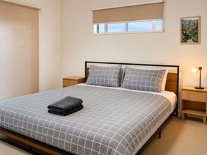 Ein ordentlich bezogenes Bett mit kariertem Überwurf und Kissen, eine Nachttischlampe, ein Nachttisch und ein Fenster mit Blick auf eine Landschaft.