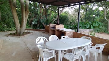 An outdoor patio with a round table and white plastic chairs, surrounded by a brick wall and covered by a metal roof.