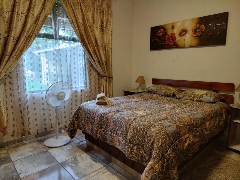 A bedroom with a patterned bedspread, a standing fan, a nightstand with a lamp, and a wall-mounted artwork.